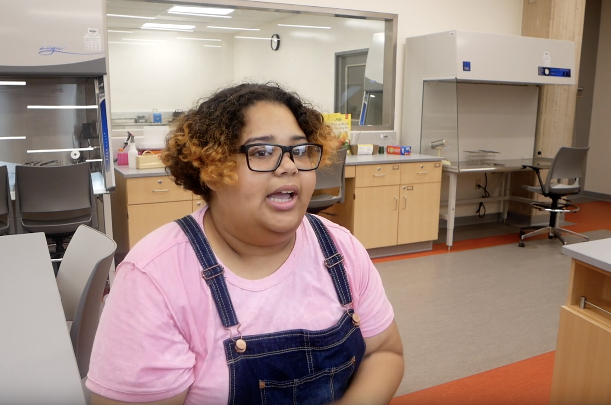 A photo of Kaishla Cabrera speaking in a lab. She is wearing a pink t-shirt and blue overalls, and glasses.